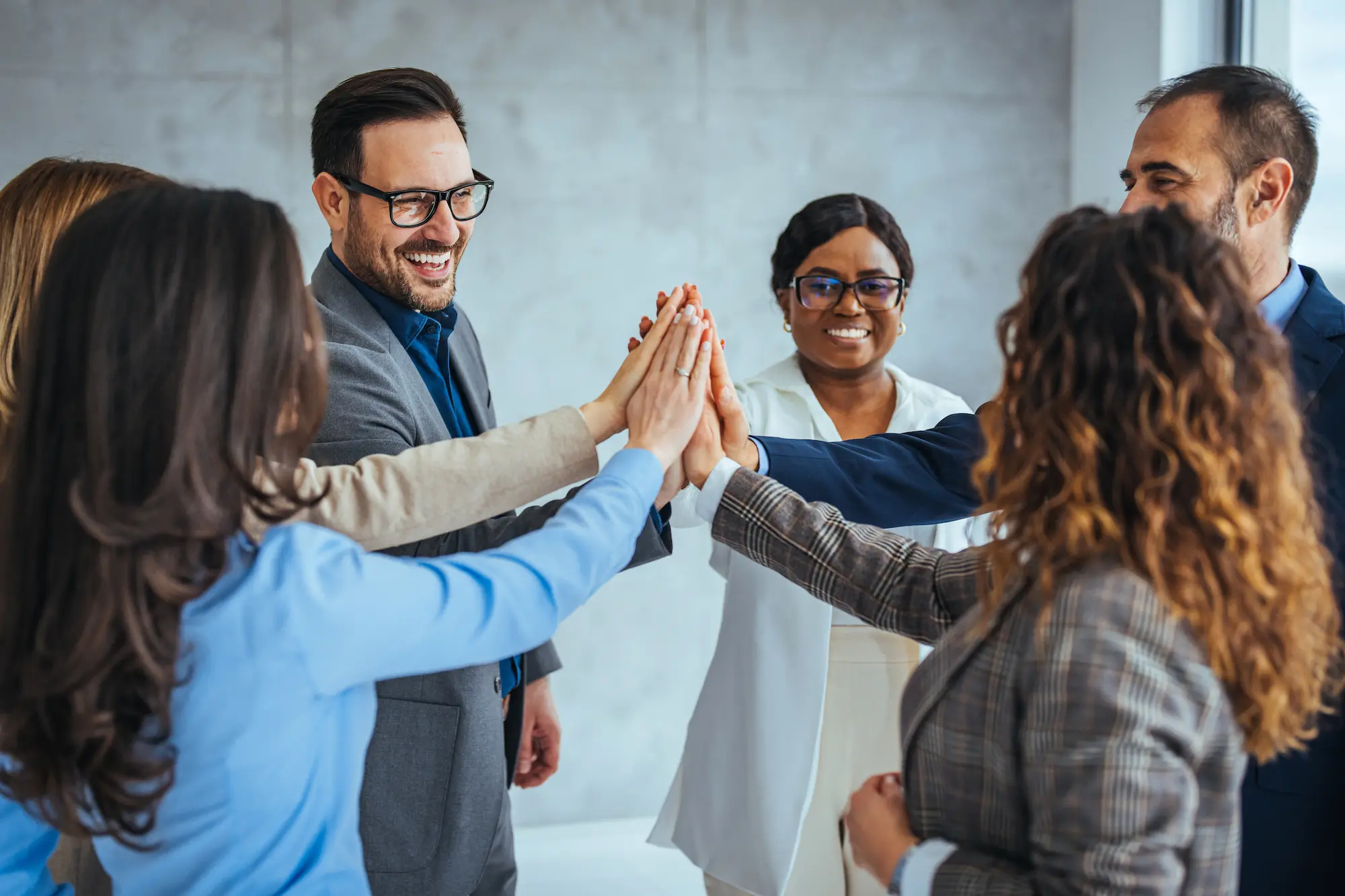 Six professionally dressed colleagues stand in a circle, smiling and joining hands for a group high-five, symbolizing teamwork and collaboration in a bright office setting.