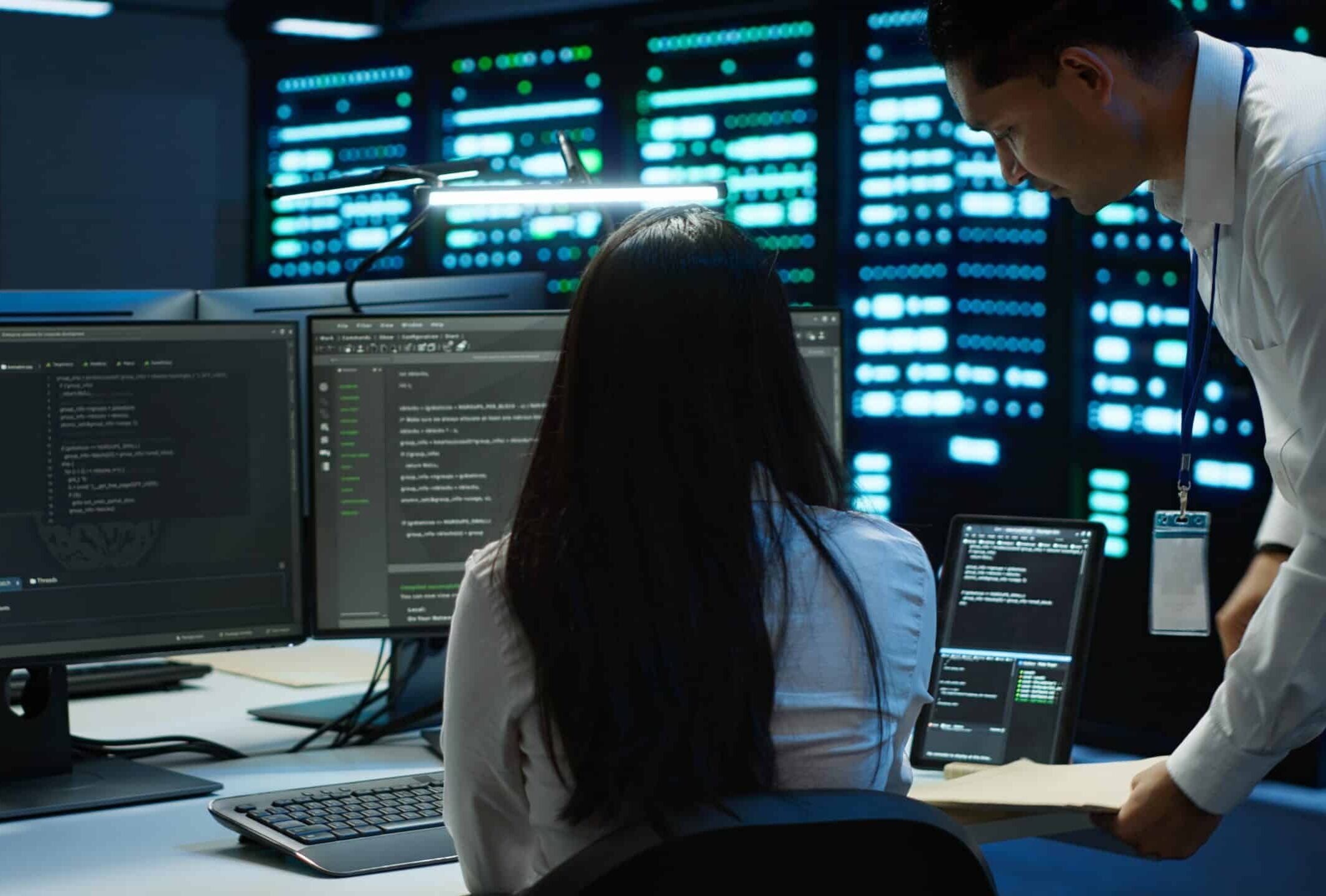 Two people in a dark server room work at a desk with multiple monitors displaying code. One sits facing the screens while the other stands nearby, holding a folder. Servers with blue lights are visible in the background.