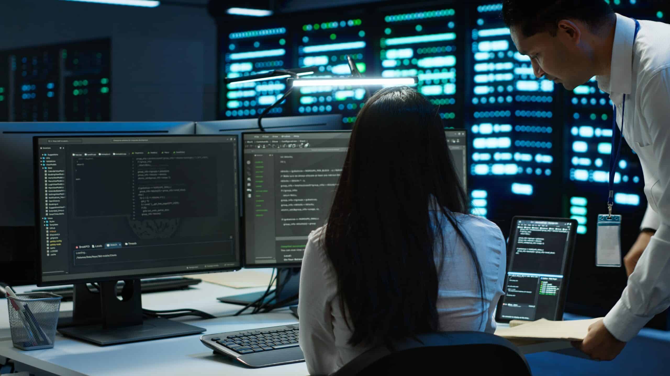 Two people in a dark server room work at a desk with multiple monitors displaying code. One sits facing the screens while the other stands nearby, holding a folder. Servers with blue lights are visible in the background.