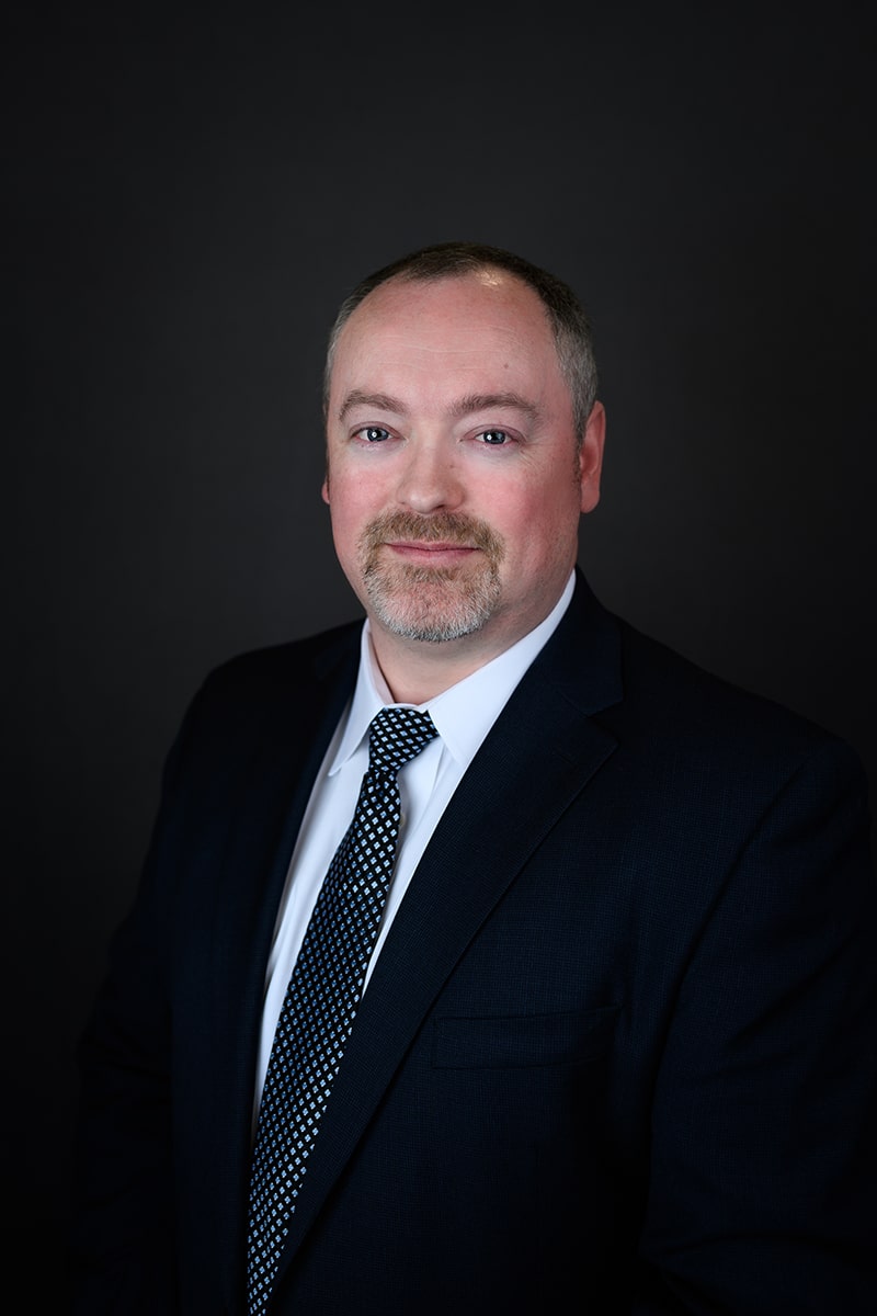A man in a dark suit, white shirt, and patterned tie poses against a plain dark background. He has short, light hair, a trimmed beard, and is looking at the camera with a slight smile.