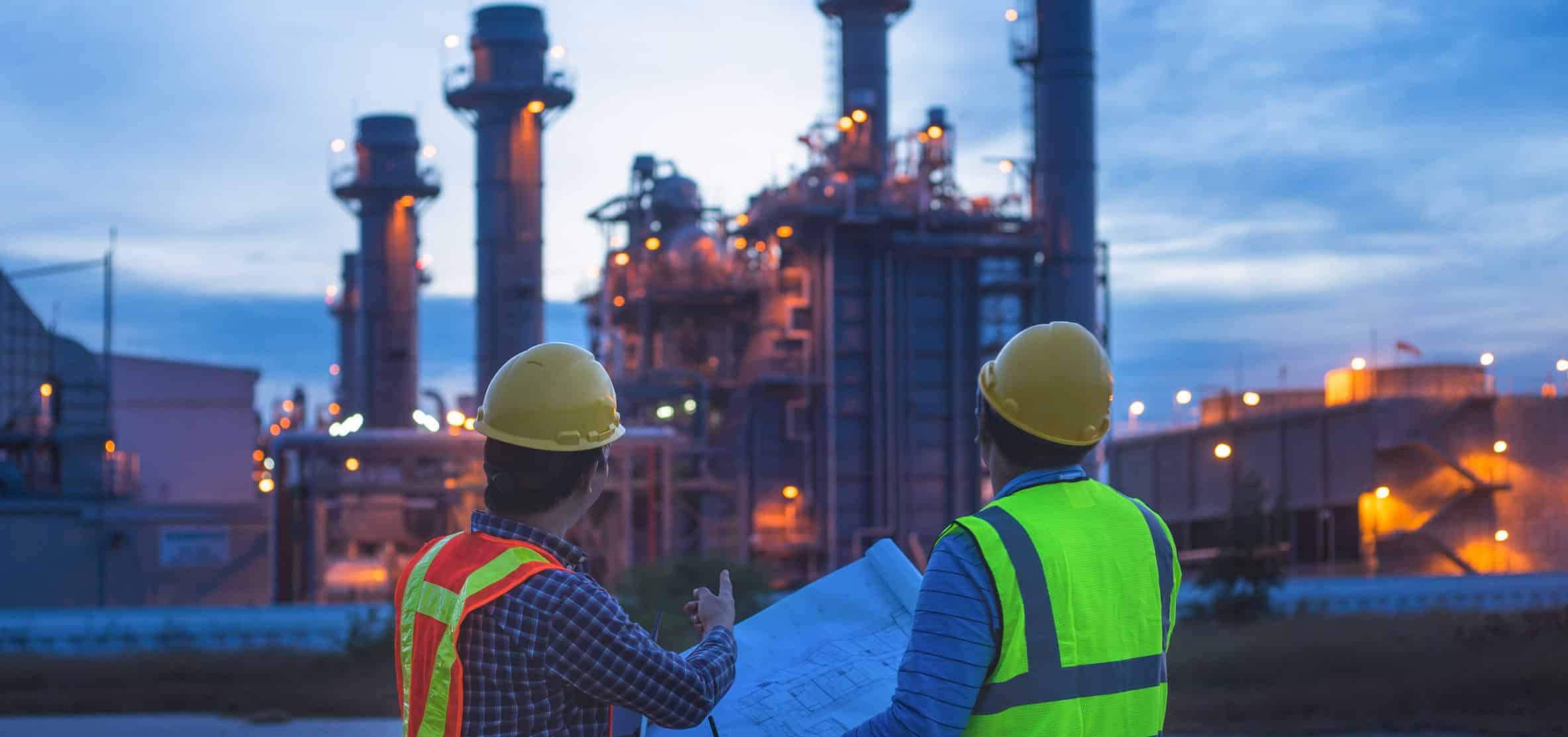 Two workers wearing safety helmets and reflective vests stand outside at dusk, looking at an industrial facility with towers and lights, while one holds blueprints or construction plans.
