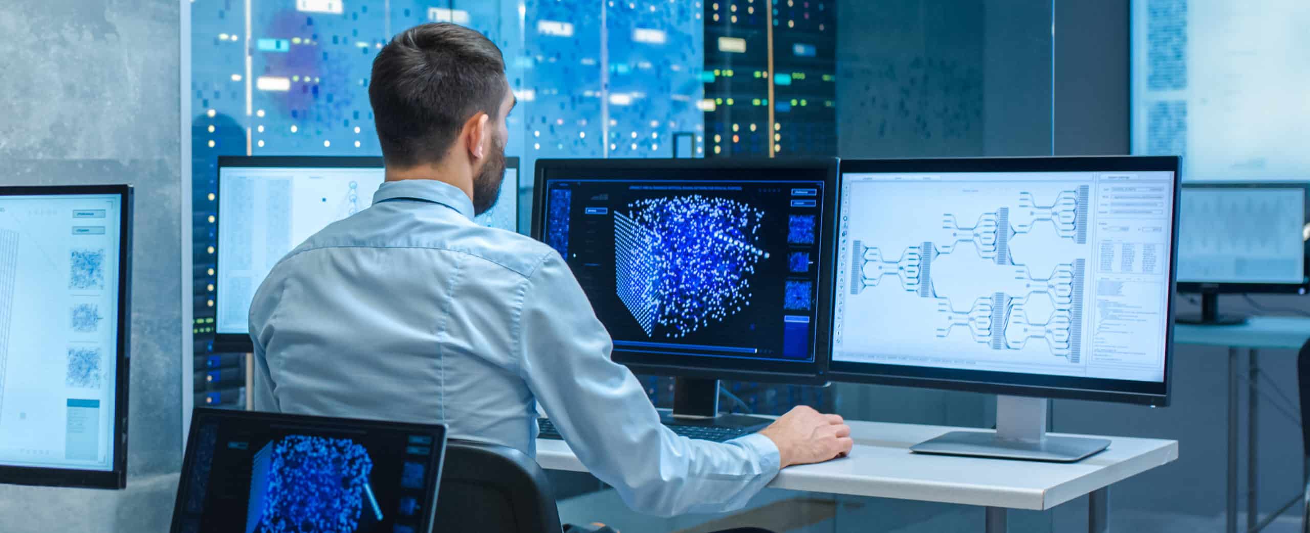 A man in a light blue shirt sits at a desk with multiple computer monitors displaying data visualizations and graphs in a modern technology control room.