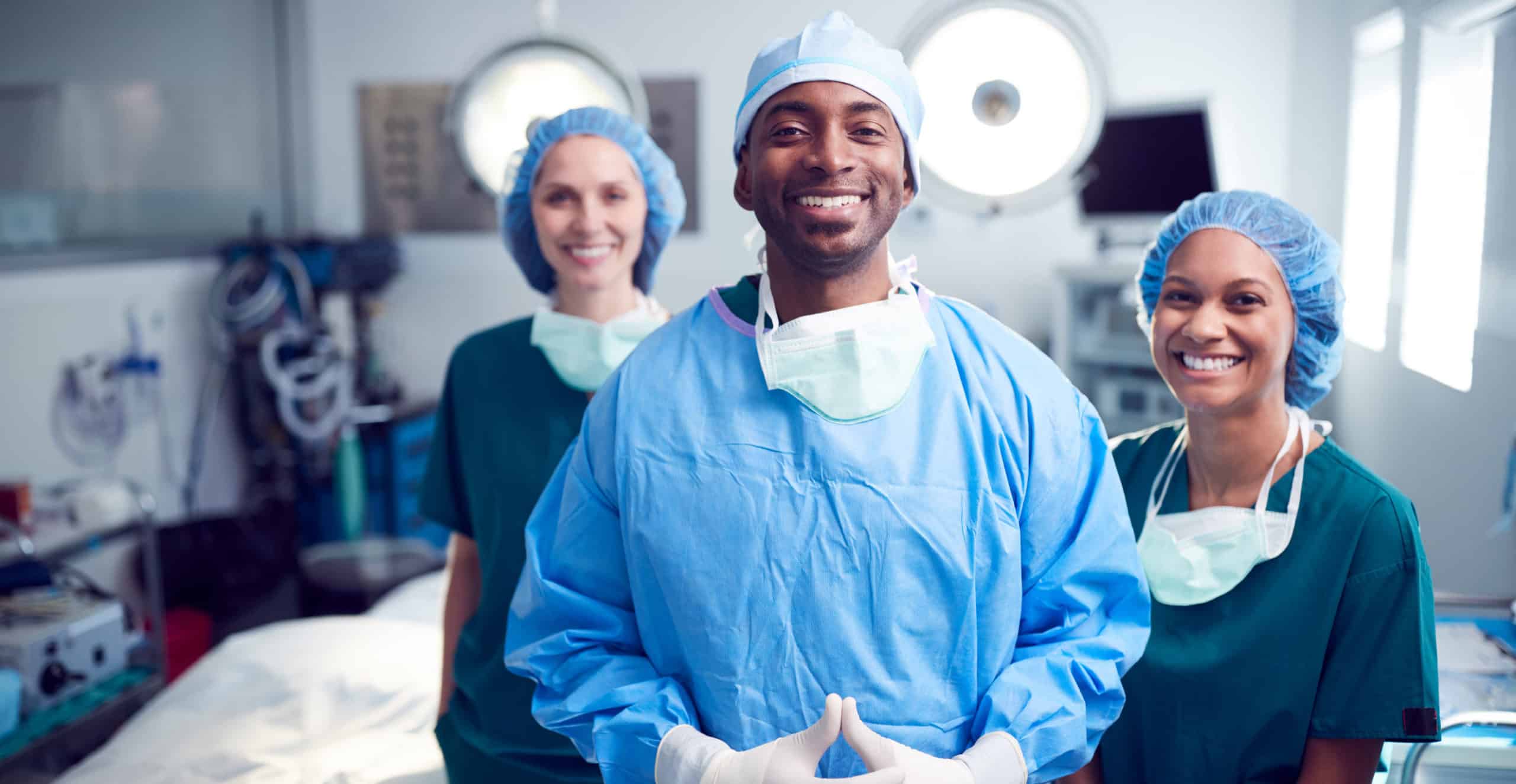 Three smiling medical professionals in surgical scrubs, masks, and caps stand together in an operating room, with one person in the foreground and two in the background.