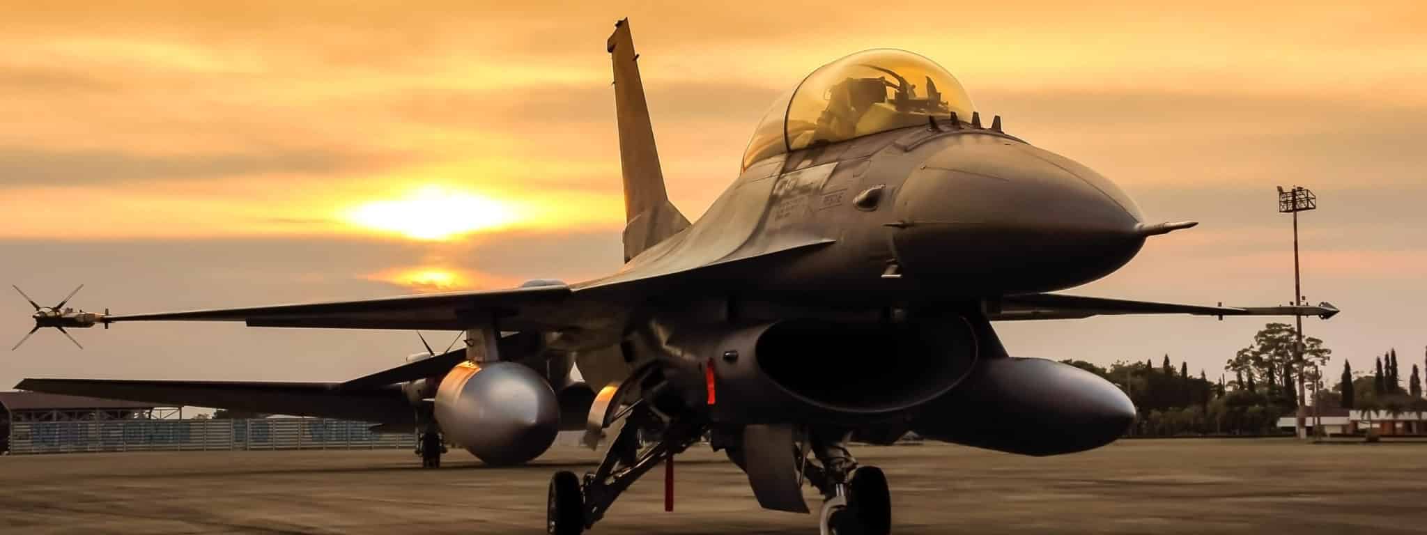 A fighter jet is parked on a runway at sunset, with the sky glowing orange and silhouettes of trees and lights in the background.