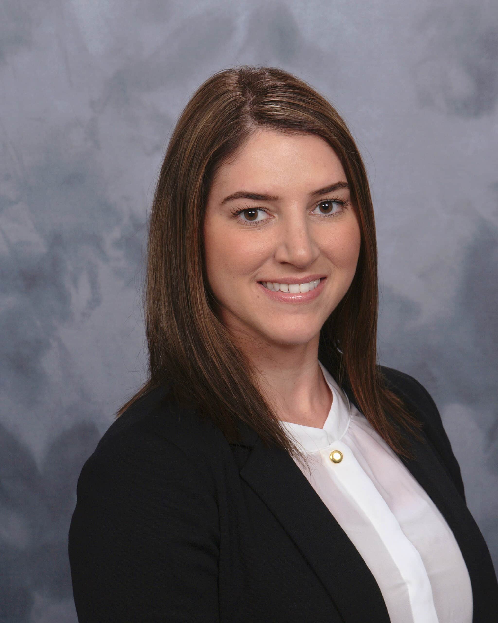 A woman with straight brown hair, wearing a black blazer over a white blouse, smiles at the camera in front of a gray mottled background.