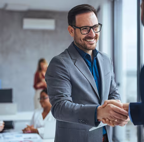 A man in a gray suit and glasses smiles while shaking hands with another person in a modern office setting. Other colleagues are working in the background.