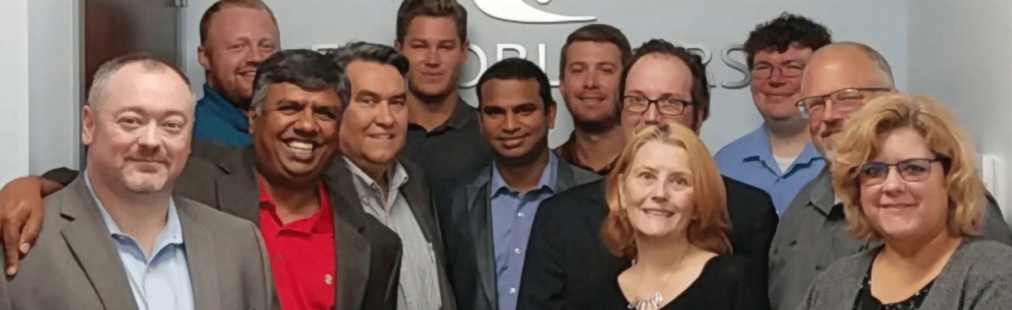 A group of twelve professionally dressed people stand closely together indoors, smiling at the camera in front of a wall with a partially visible logo.