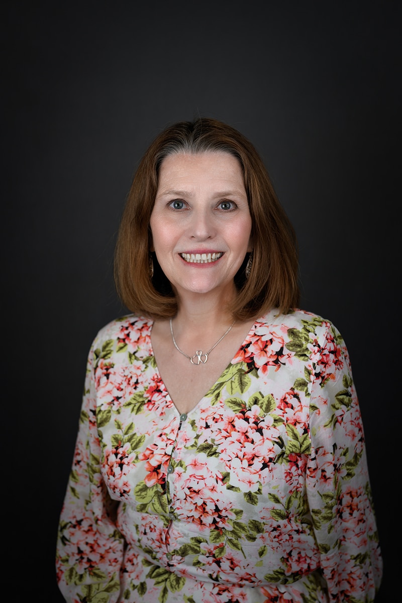 A woman with shoulder-length brown hair smiles at the camera, wearing a floral-patterned top with pink and green flowers and a silver necklace, set against a dark plain background.