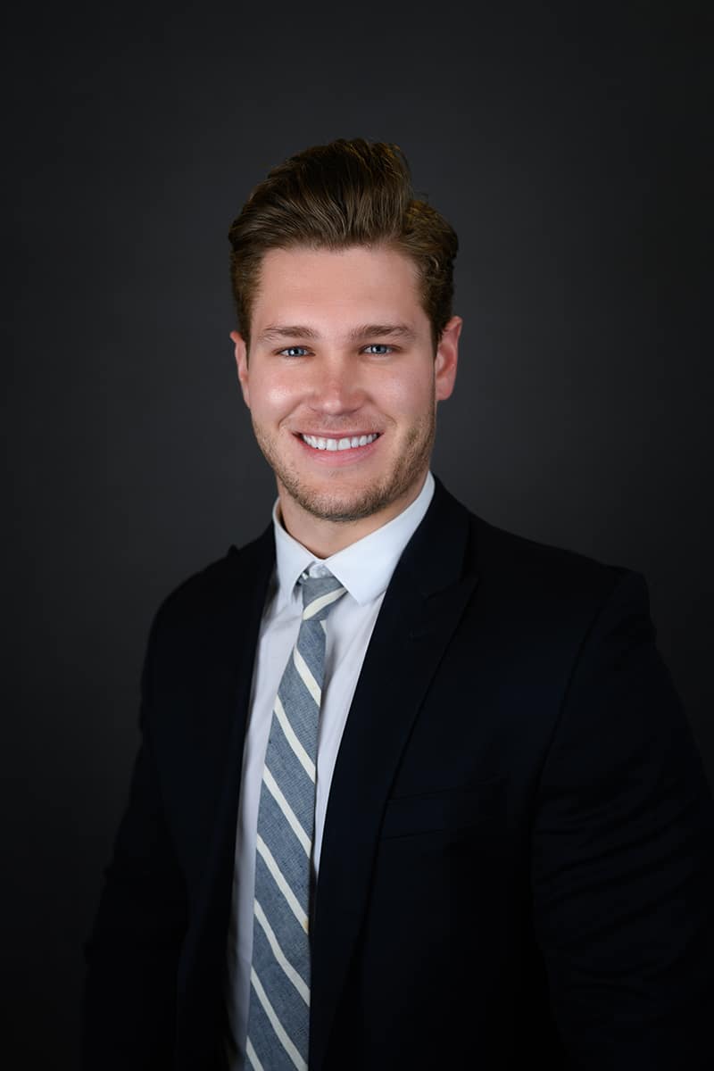 A young man with light brown hair is smiling and wearing a dark suit, white shirt, and striped tie, standing against a plain dark background.