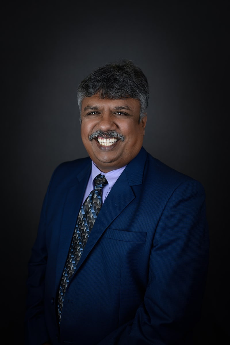 A man with gray hair and a mustache, wearing a blue suit, patterned tie, and lavender shirt, smiles broadly against a dark background.