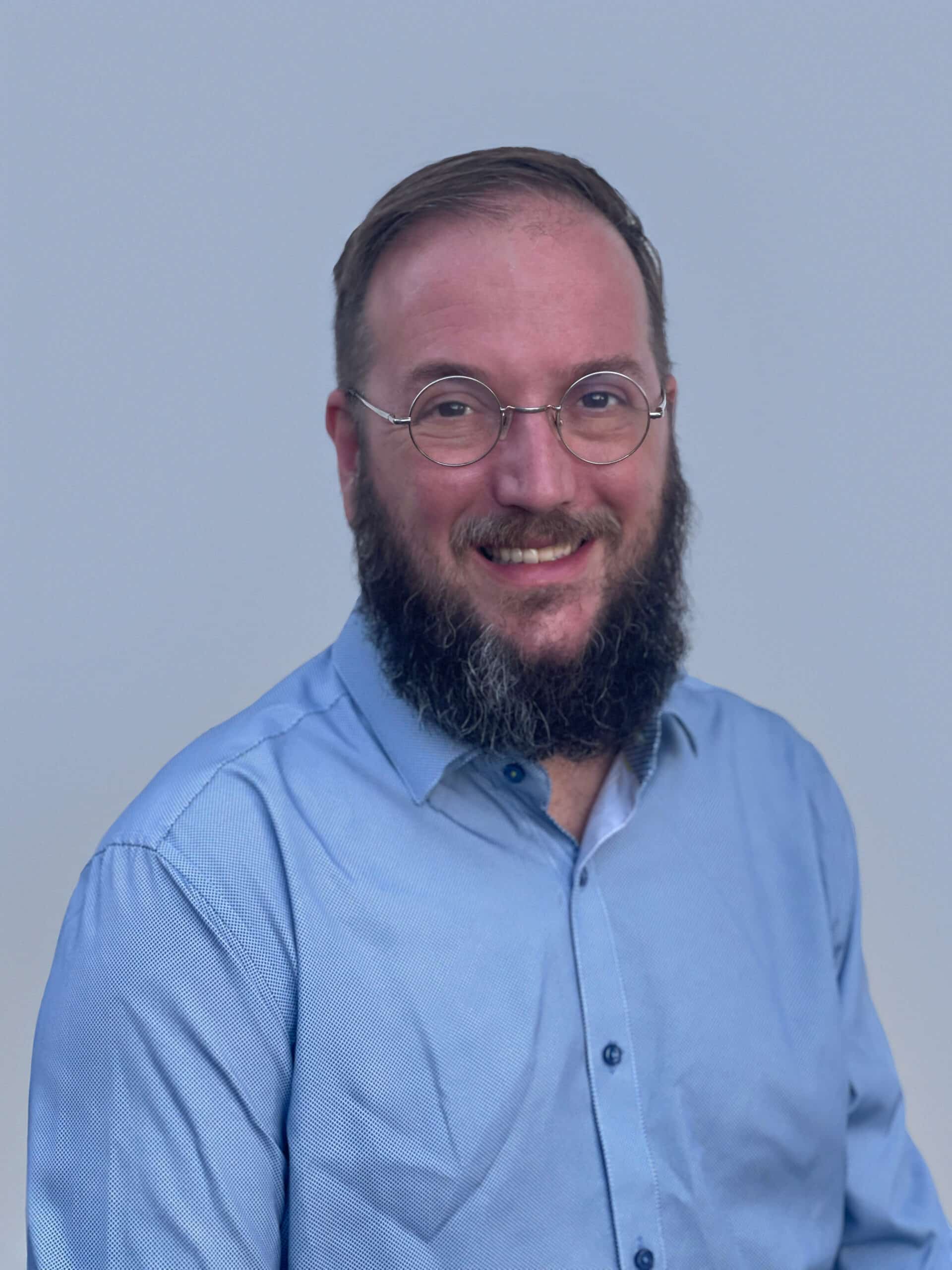 A man with glasses and a beard, wearing a light blue button-up shirt, smiles while facing the camera against a plain light background.