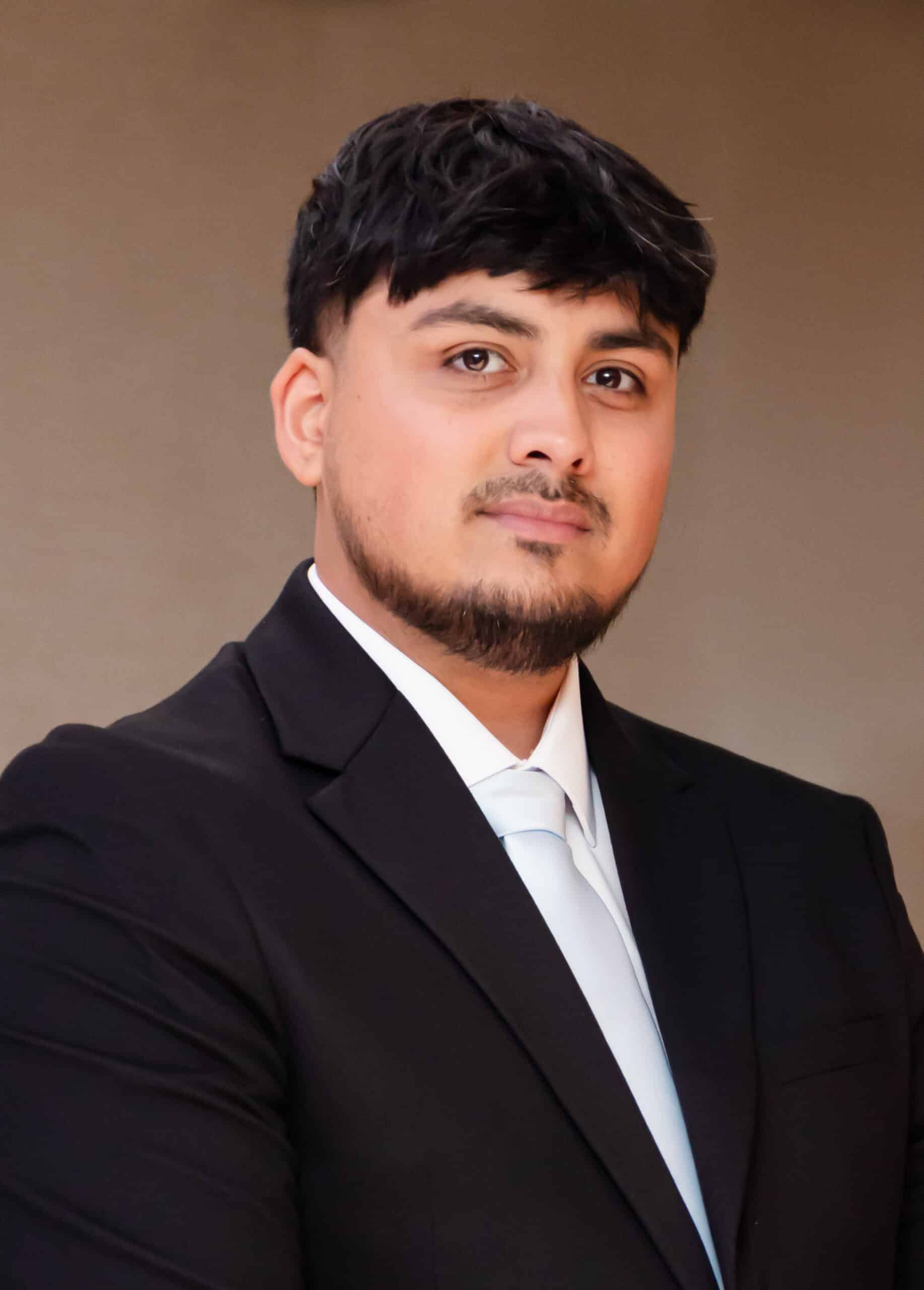 A man with short black hair and a beard is wearing a black suit, white shirt, and light blue tie, posing against a plain, neutral background. He is looking confidently at the camera.