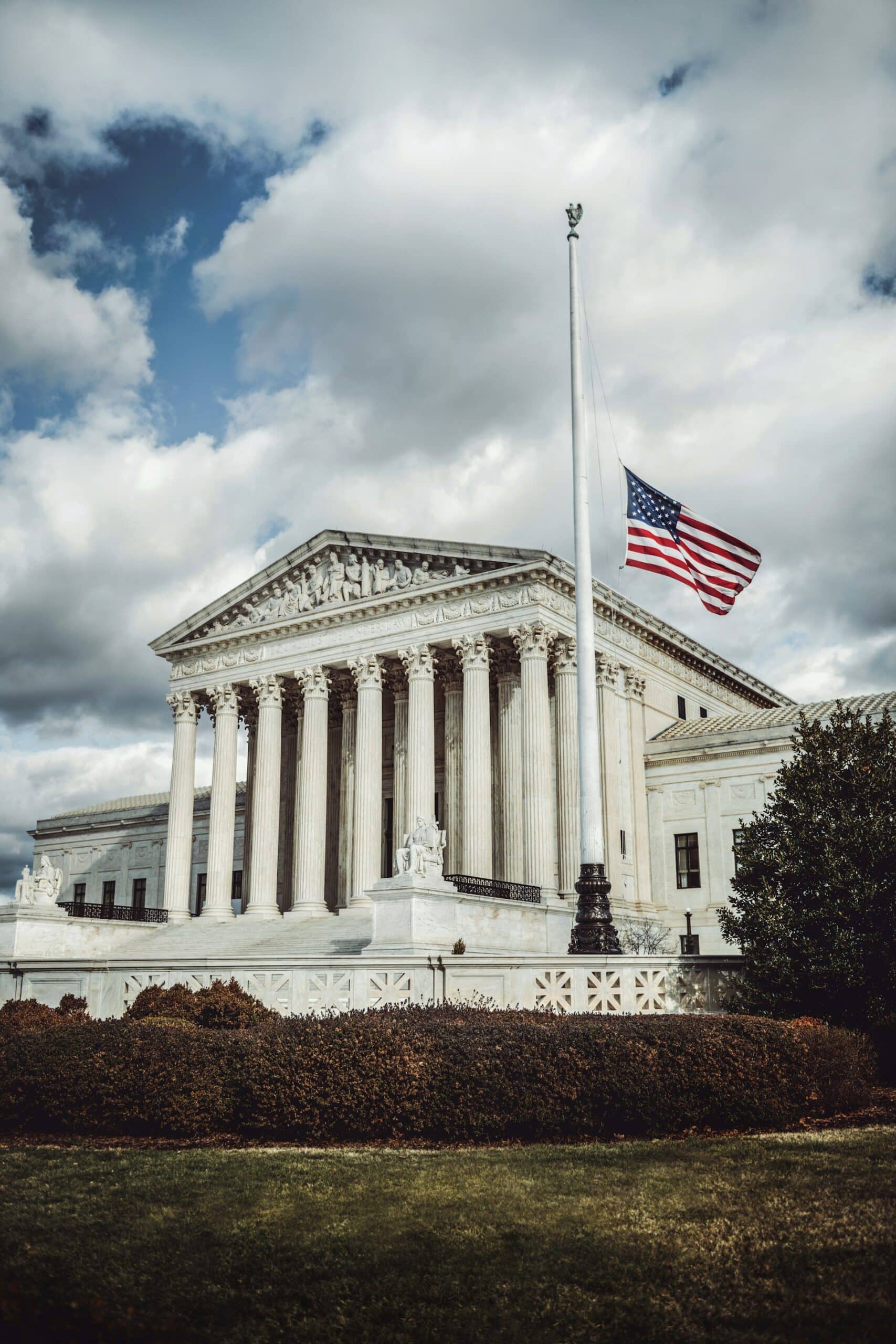 The American flag flies at half-staff in front of the U.S. Supreme Court building, which features tall columns and classical architecture under a cloudy sky.