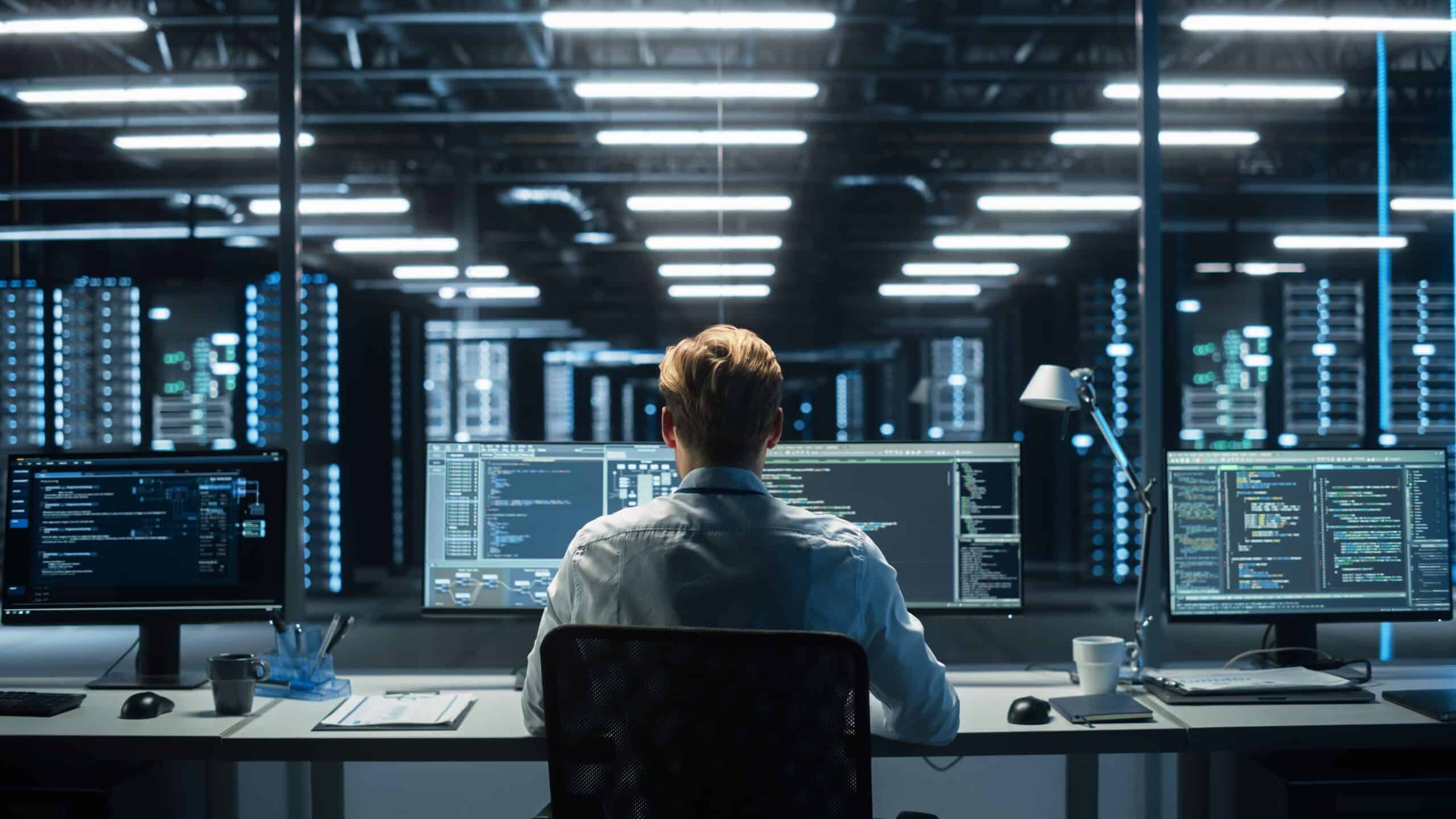 A person sits at a desk with multiple computer monitors displaying code and system data in a large, modern server room with rows of illuminated server racks.