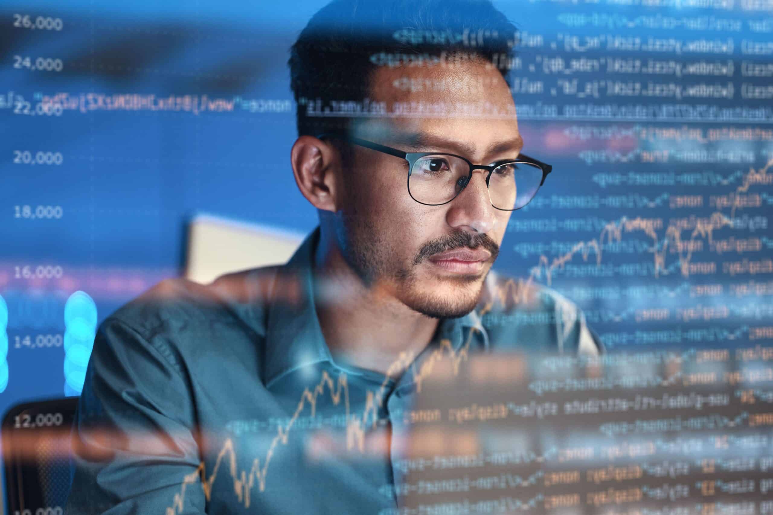 A man wearing glasses focuses on a computer screen displaying lines of code and a financial graph, suggesting data analysis or programming in a technology or finance setting.
