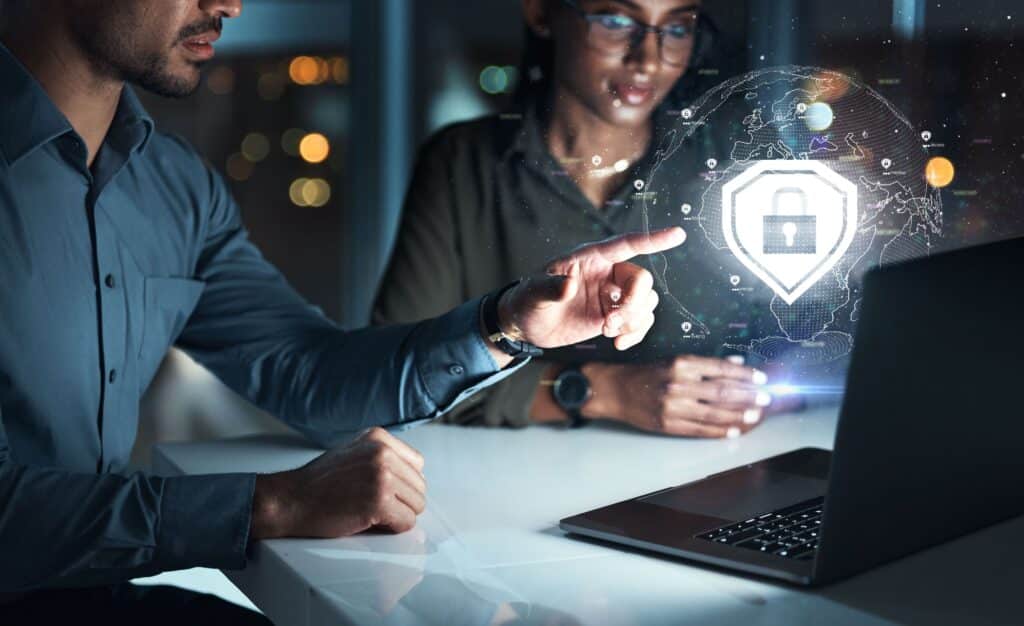 Two people sit at a desk by a laptop, one pointing at a floating holographic padlock symbol, representing cybersecurity or data protection, with city lights visible through the window in the background.