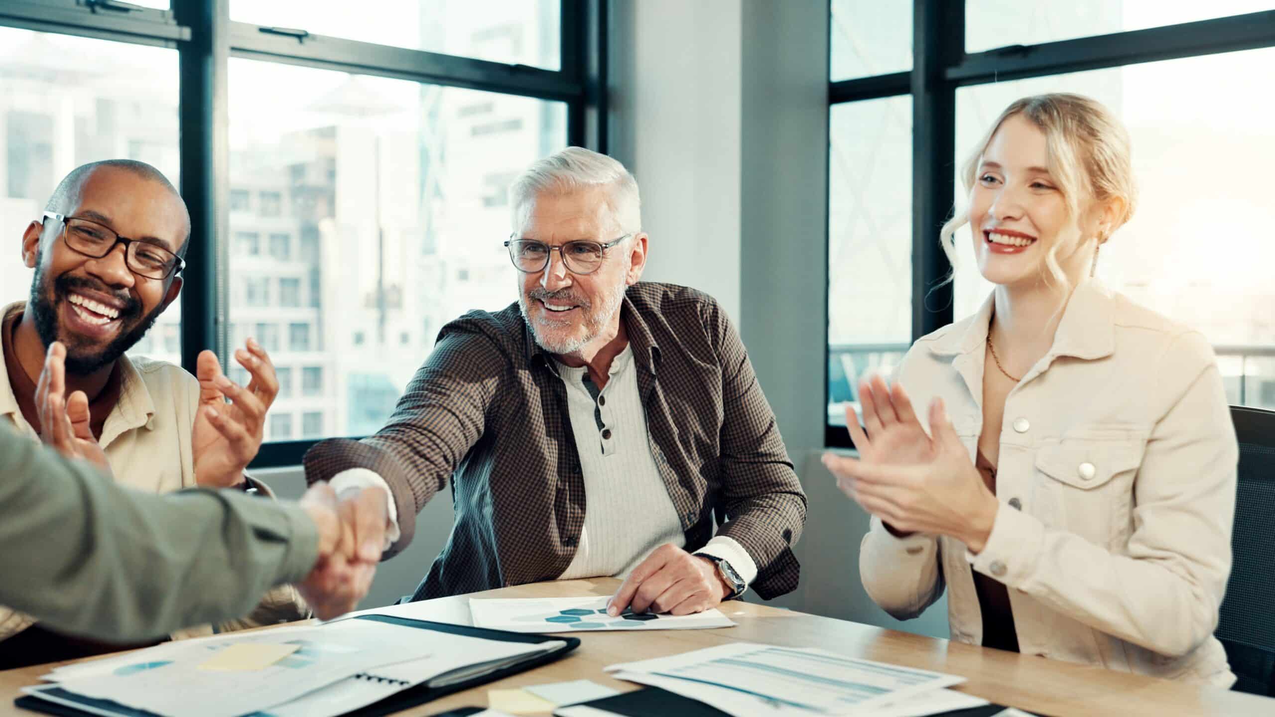 Three businesspeople sit at a table in a modern office, smiling and clapping while one man shakes hands with another person across the table, celebrating a successful meeting or agreement.